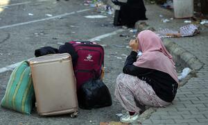 A woman wearing a hijab sits on a curb in Beirut next to her luggage, appearing distressed, amidst debris on the street following a mass evacuation warning.