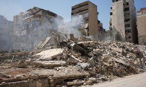 A street in the Bashura neighborhood of Beirut, Lebanon, covered in rubble and debris from destroyed buildings following airstrikes, with smoke rising from the wreckage.