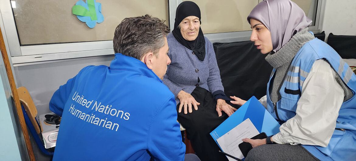 Tom Fletcher, coordinateur des secours d'urgence de l'ONU, vêtu d'une veste humanitaire bleue des Nations Unies, s'assoit et parle avec Setta Ayyoub, une femme âgée en hijab noir qui a été déplacée par le conflit. Ils sont à l'école publique Omar Onsi, un refuge collectif à Beyrouth, accompagnés d'une autre femme en hijab et gilet bleu tenant un dossier.