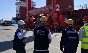 WHO and UNOPS officials in safety vests and hard hats at a port, overseeing the loading of humanitarian aid onto a cargo ship for delivery to Gaza.