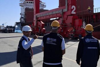 WHO and UNOPS officials in safety vests and hard hats at a port, overseeing the loading of humanitarian aid onto a cargo ship for delivery to Gaza.