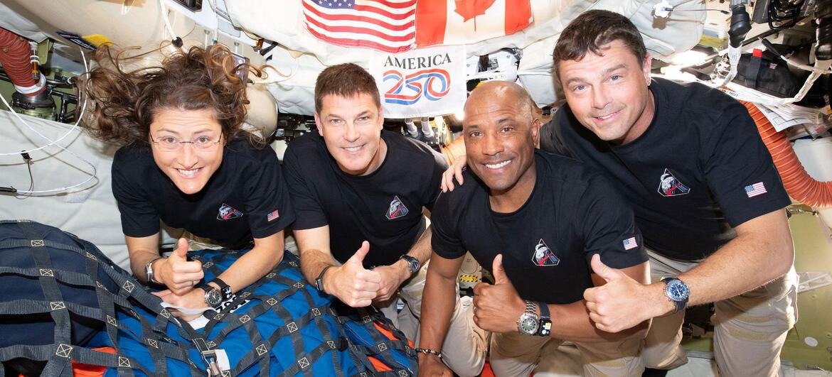 NASA Artemis II astronauts Reid Wiseman, Victor Glover, Christina Koch, and Jeremy Hansen pose with thumbs up inside the Orion spacecraft, with American and Canadian flags and an America 250 sign in the background.