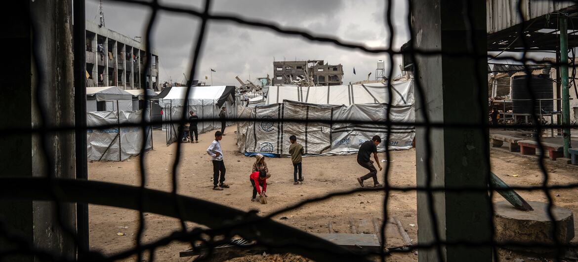 Des enfants jouent dans une cour d'un site de déplacement à Jabalia, à Gaza, vue à travers une clôture en fil de fer avec des tentes et des bâtiments détruits en arrière-plan.