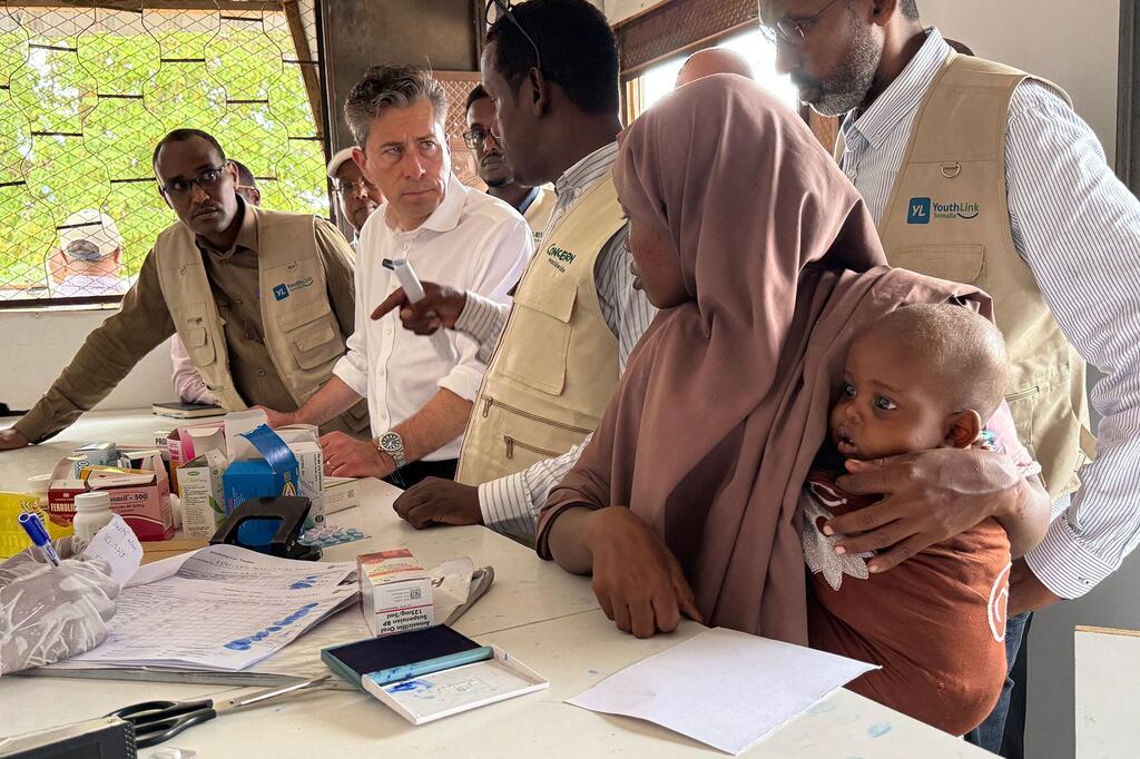 Naibu Katibu Mkuu wa Umoja wa Mataifa wa Masuala ya Kibinadamu Tom Fletcher anatembelea kliniki ya afya huko Somalia, ambapo anazungumza na mama anayemshika mtoto wake aliye na utapiamlo wakati wa uchunguzi wa matibabu.