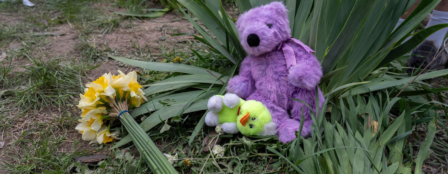 Un ours en peluche violet et un bouquet de jonquilles jaunes sont déposés sur la pelouse en guise d'hommage. Un petit canard en peluche vert est niché devant l'ours.