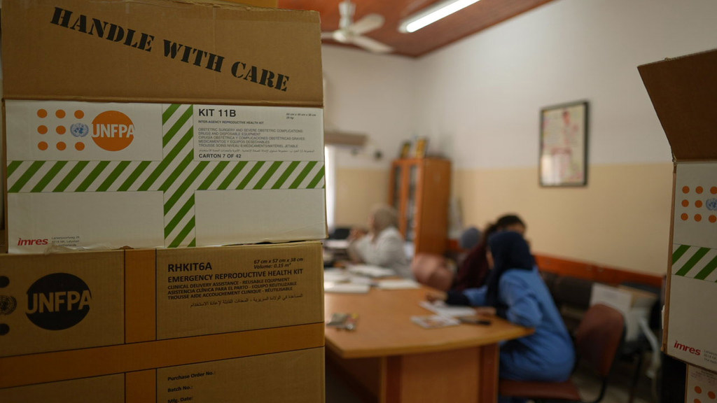 तस्वीर में आगे की ओर UNFPA की आपातकालीन प्रजनन स्वास्थ्य किटें रखी हुई हैं, जबकि पृष्ठभूमि में अस्पताल के माहौल में चिकित्सा कर्मचारी एक डेस्क पर काम कर रहे हैं।