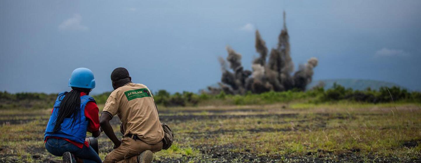 A UN peacekeeper in a blue helmet and an AFRILAN demining specialist crouch in a field watching a controlled mine explosion in the background.