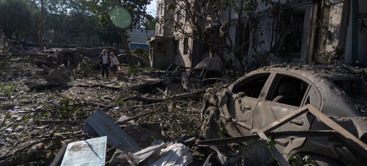 A destroyed car and rubble in a damaged building in Kyiv, Ukraine, with a woman and a child walking through the debris.