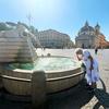 Une femme âgée en robe blanche se tient à côté d'une grande fontaine en pierre sur une place d'une ville européenne, se touchant le front à cause de la chaleur.