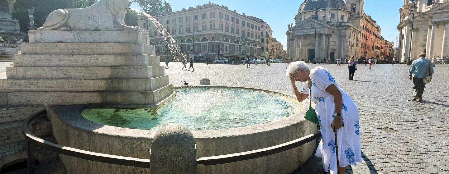Une femme âgée en robe blanche se tient à côté d'une grande fontaine en pierre sur une place d'une ville européenne, se touchant le front à cause de la chaleur.