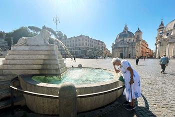 Une femme âgée en robe blanche se tient à côté d'une grande fontaine en pierre sur une place d'une ville européenne, se touchant le front à cause de la chaleur.
