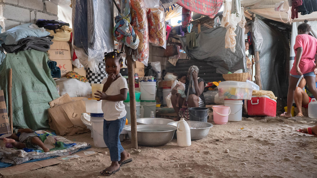 Una joven haitiana se encuentra dentro de una carpa abarrotada en Puerto Príncipe, sosteniendo una taza, mientras otras personas desplazadas descansan en el fondo.