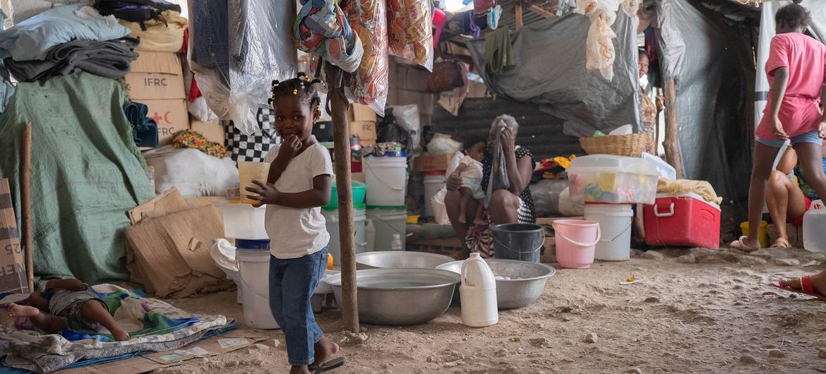 A young Haitian girl stands inside a crowded tent shelter in Port au Prince, holding a cup, while other displaced people rest in the background.