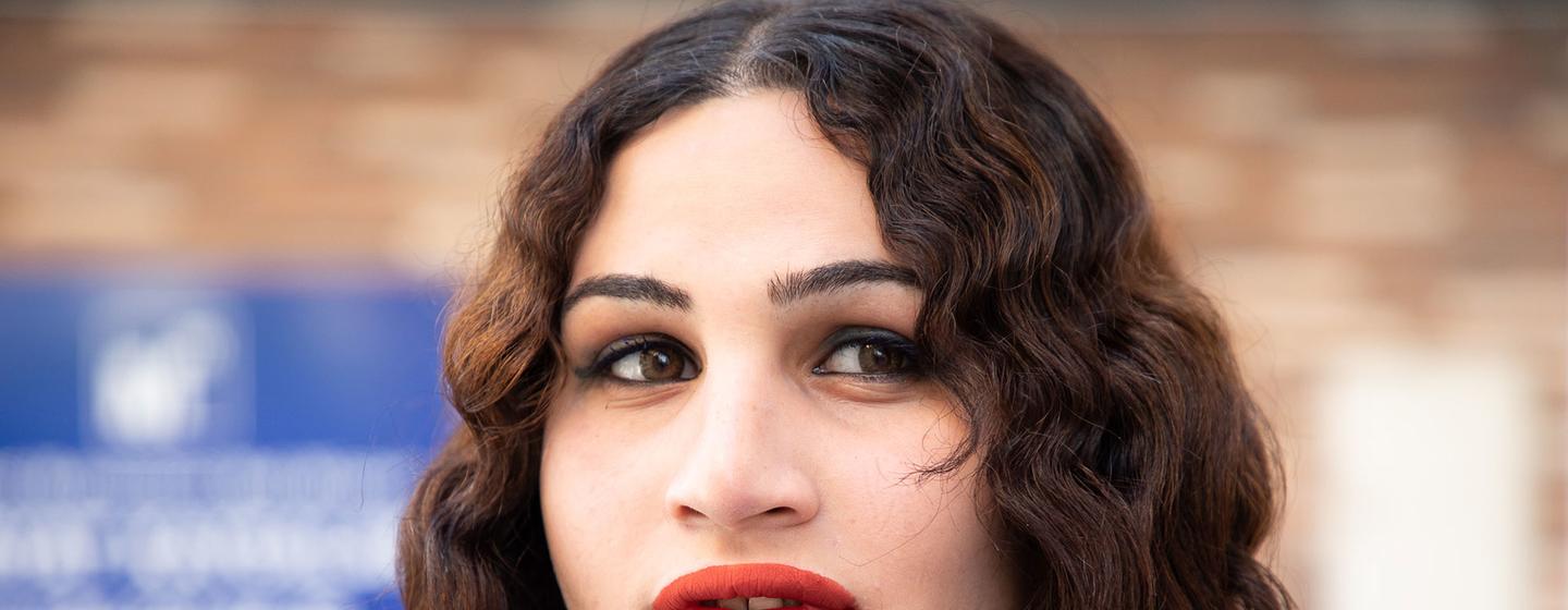 Retrato de una joven con cabello oscuro y rizado y lápiz labial rojo, con el rostro reflejado en una pared de ladrillos.