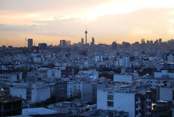 A panoramic view of the city of Tehran, Iran, at sunset. The skyline features numerous buildings and a prominent tower against a hazy, golden sky.