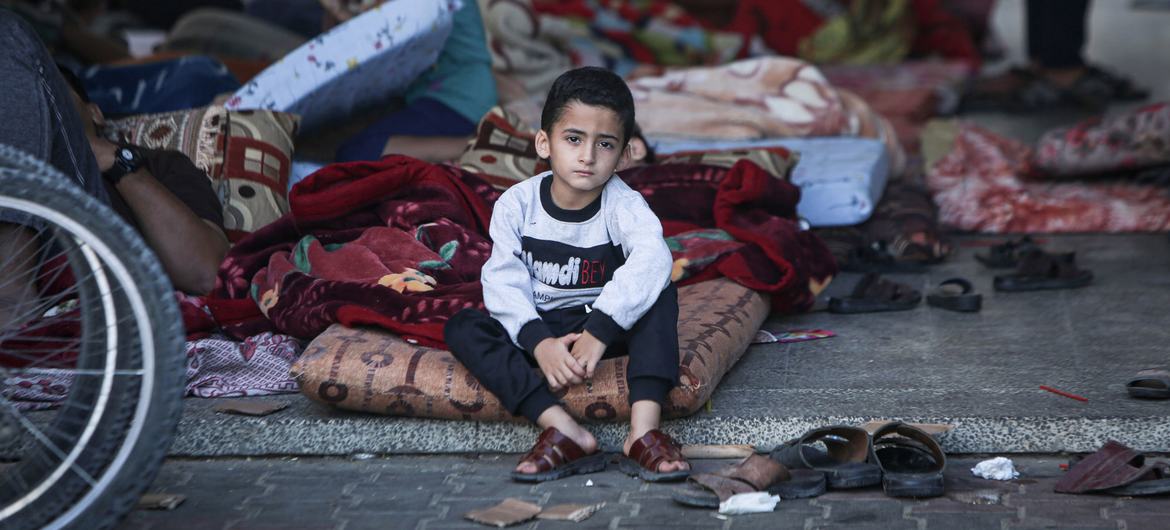 A five-year-old boy sits on his mattress in a shelter courtyard surrounded by several hundred other displaced people.