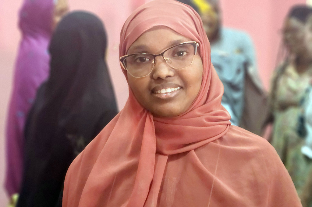 Una mujer sonriente con hiyab rosa y gafas en Yibuti, con otras personas al fondo.
