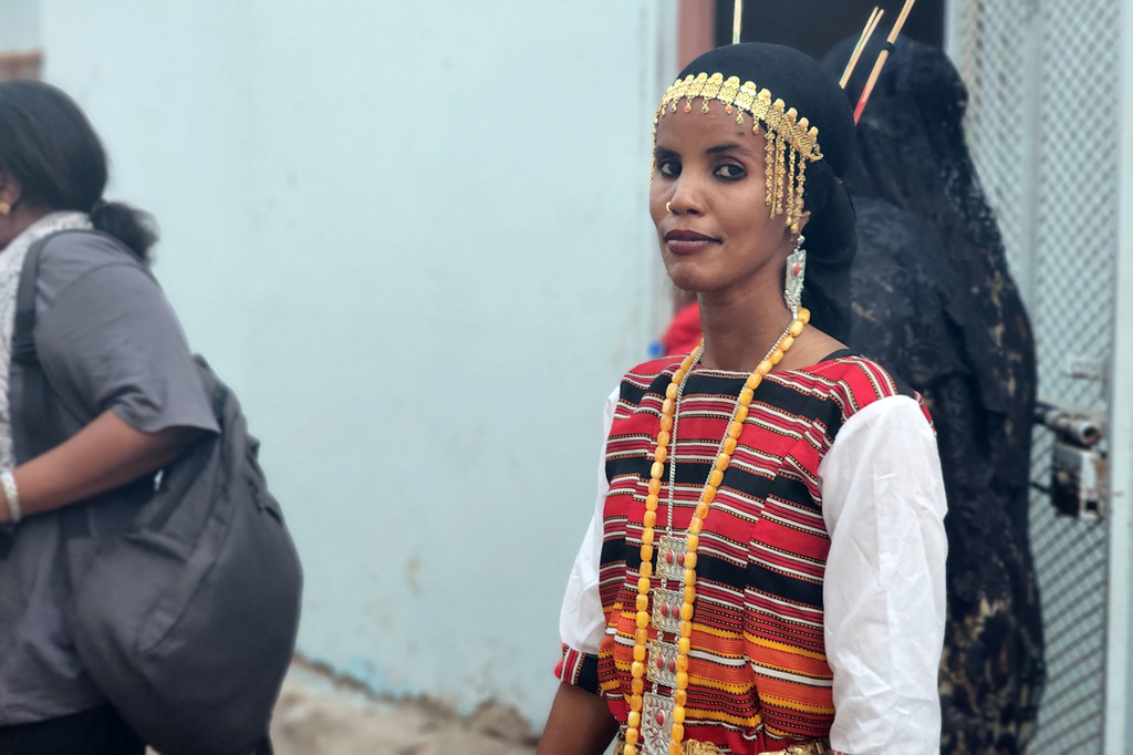 Una mujer vestida con el traje tradicional de Yibuti, con joyas de oro y un collar de cuentas, permanece de pie mirando hacia la cámara.