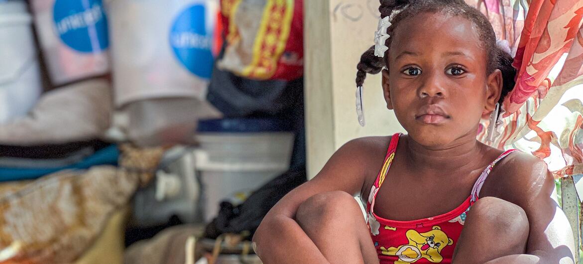 A young girl, age 9, sits quietly in a crowded public shelter in Haiti, where her family has sought refuge from armed group violence. She is surrounded by basic supplies, including UNICEF hygiene kits, highlighting the organization's efforts to combat waterborne diseases in displacement sites.