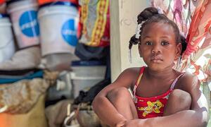 A young girl, age 9, sits quietly in a crowded public shelter in Haiti, where her family has sought refuge from armed group violence. She is surrounded by basic supplies, including UNICEF hygiene kits, highlighting the organization's efforts to combat waterborne diseases in displacement sites.
