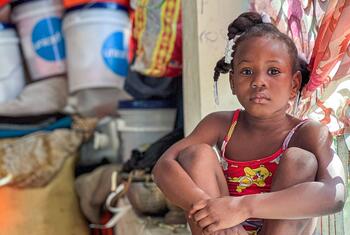 A young girl, age 9, sits quietly in a crowded public shelter in Haiti, where her family has sought refuge from armed group violence. She is surrounded by basic supplies, including UNICEF hygiene kits, highlighting the organization's efforts to combat waterborne diseases in displacement sites.