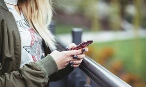 A woman with blonde hair and dark nail polish is using a smartphone while leaning on a railing outdoors.