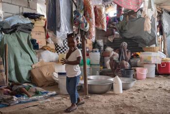 Une jeune fille se tient dans un camp encombré et improvisé en Haïti, entouré de tentes et de seaux, soulignant les conditions de vie difficiles auxquelles sont confrontées les familles déplacées.