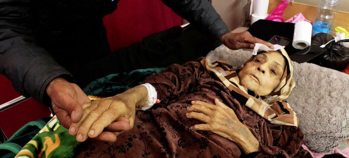 An elderly cancer patient lies in bed in Gaza, receiving care from a person who is gently holding her hand and wiping her forehead.