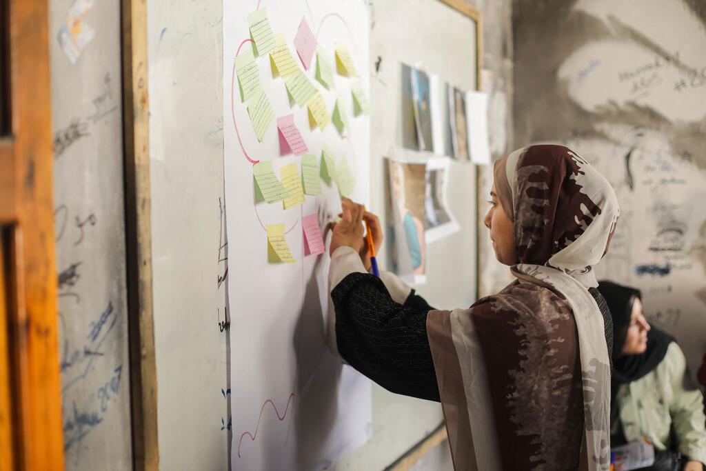 Mwanamke mmoja huko Gaza, akiwa amevaa hijabu, anashiriki kikamilifu katika kikao cha kupanga jamii kwa kuandika kwenye ubao mkubwa uliofunikwa na noti za rangi. Mwanamke mwingine anaonekana nyuma, akiashiria juhudi za pamoja za kukabiliana na changamoto za mitaa.