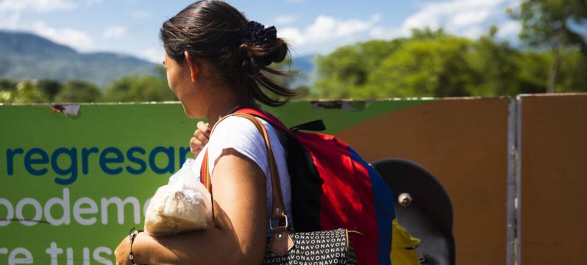 A Latin American woman with a backpack and bags walks outdoors with mountains in the background.