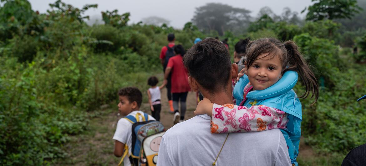 A Venezuelan migrant father carries his young daughter on his shoulders as they walk through a jungle path with other migrants ahead.
