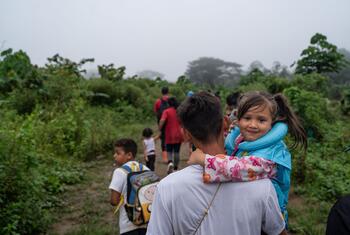 A Venezuelan migrant father carries his young daughter on his shoulders as they walk through a jungle path with other migrants ahead.