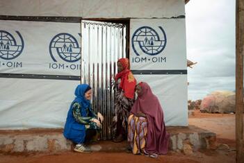 An IOM aid worker crouches to speak with two displaced women wearing headscarves at the entrance of an IOM field office in a refugee camp in Sudan.