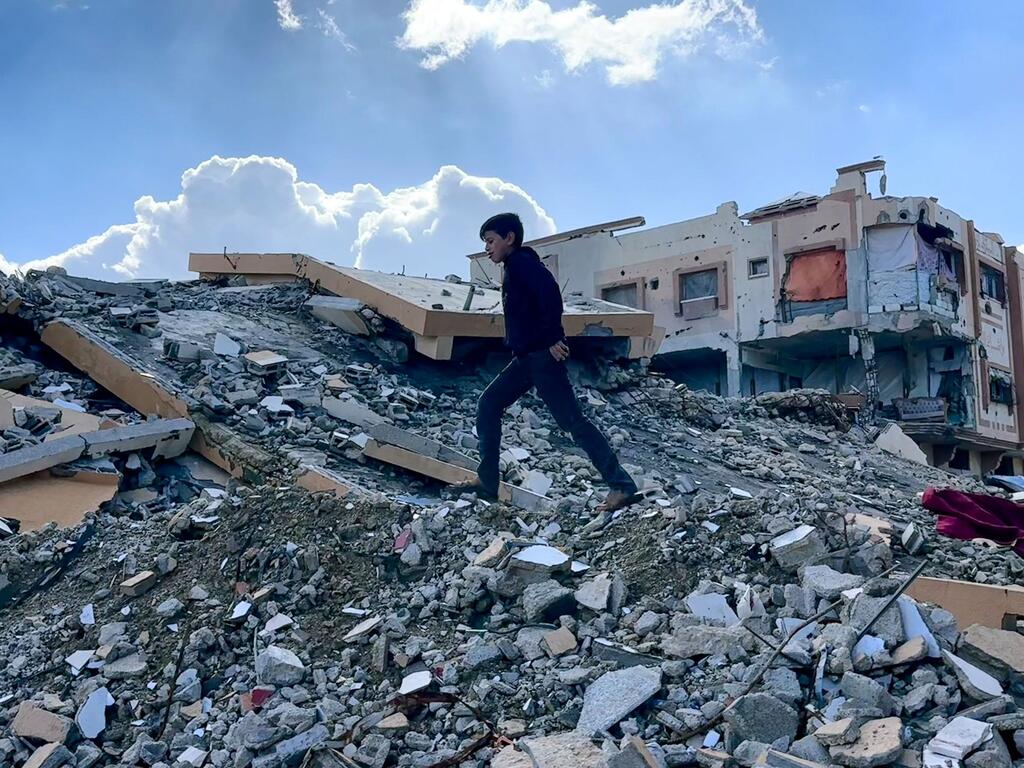 Un garçon palestinien marche sur les décombres de maisons détruites à Gaza, avec des bâtiments endommagés en arrière-plan.