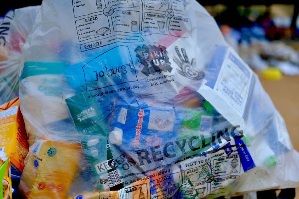 Un sac de recyclage en plastique transparent rempli de divers articles recyclables, notamment des bouteilles en plastique, des cartons et des contenants, avec un guide de recyclage de Johannesburg visible sur le sac.