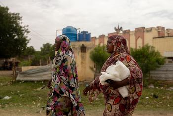 Deux femmes portant des foulards colorés marchent ensemble, l'une d'elles tenant un ballot de tissu blanc. À l'arrière-plan, on aperçoit des réservoirs d'eau et un bâtiment dans un cadre villageois.