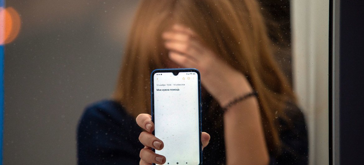 A teenage girl holds a smartphone with a text message, her face blurred, looking through a glass window.