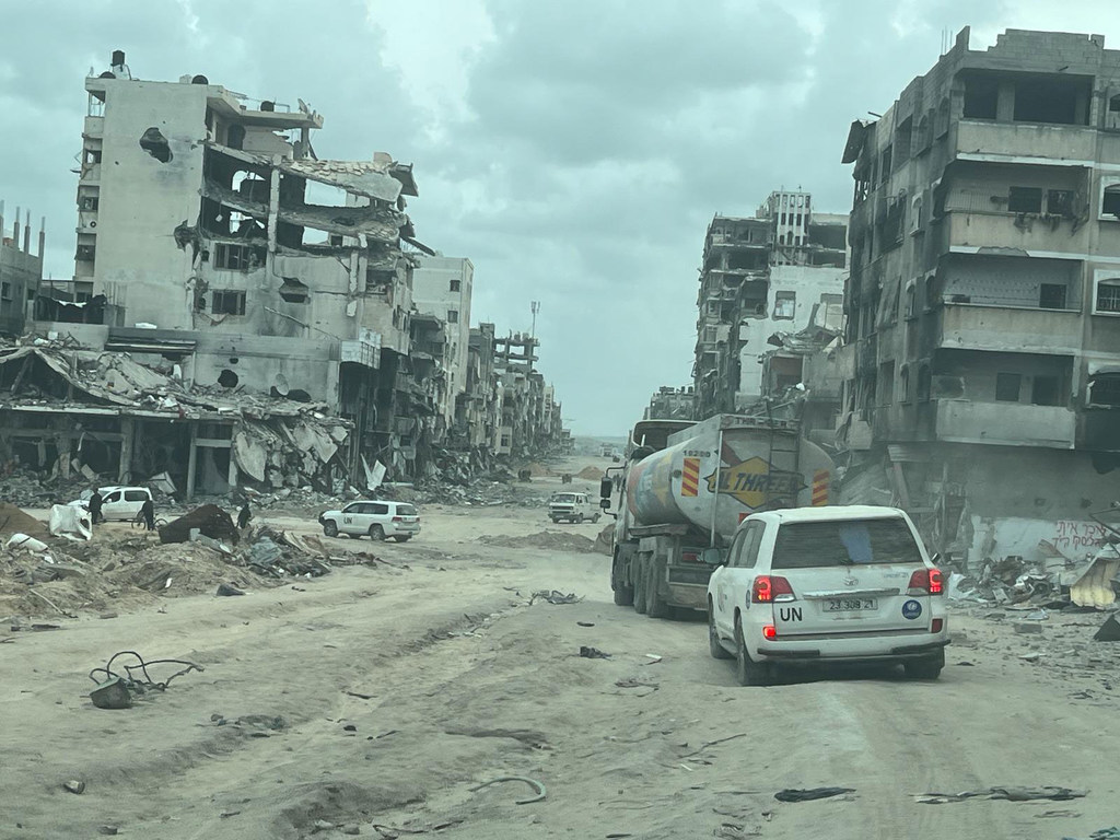 Un vehículo blanco de la ONU y un camión cisterna de combustible circulan por un camino de tierra flanqueado por edificios gravemente dañados y destruidos en el norte de Gaza.