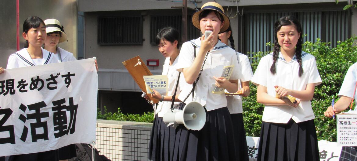 A group of Japanese high school students in uniforms are participating in a protest or demonstration, holding signs and using a megaphone to speak.