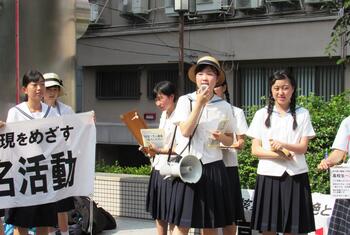 Un groupe d'élèves du secondaire japonais en uniforme participe à une manifestation ou à une manifestation, tenant des pancartes et utilisant un mégaphone pour parler.