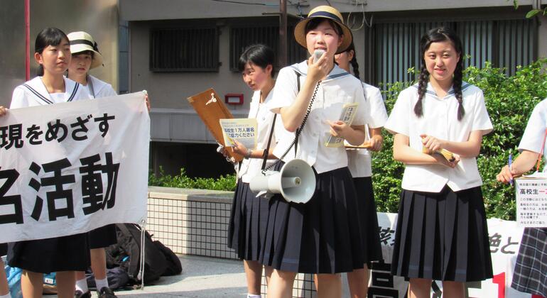Un groupe d'élèves du secondaire japonais en uniforme participe à une manifestation ou à une manifestation, tenant des pancartes et utilisant un mégaphone pour parler.