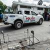 Uma caminhonete branca da polícia haitiana está estacionada em frente a um prédio em Porto Príncipe, Haiti, com uma barricada de metal em primeiro plano. Uma multidão de pessoas é visível ao fundo.