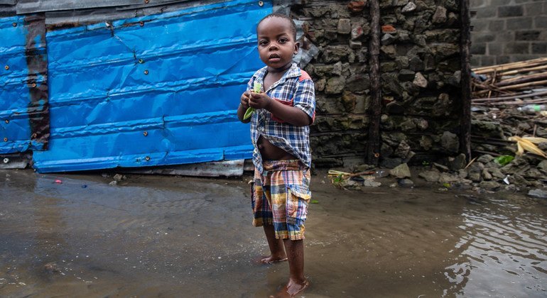 A young African boy stands barefoot in rising floodwater in front of a makeshift shelter with blue tarp and stone walls in Praia Nova, Beira, Mozambique.