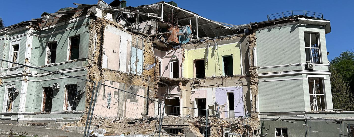 Un bâtiment de plusieurs étages gravement endommagé, avec un toit effondré et des murs intérieurs exposés, présentant des signes de dégâts de guerre, avec une clôture au premier plan et de l'herbe poussant à sa base.