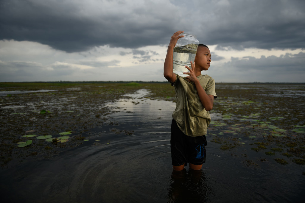Un garçon se tient debout dans un champ inondé, portant un grand récipient d'eau sur son épaule.