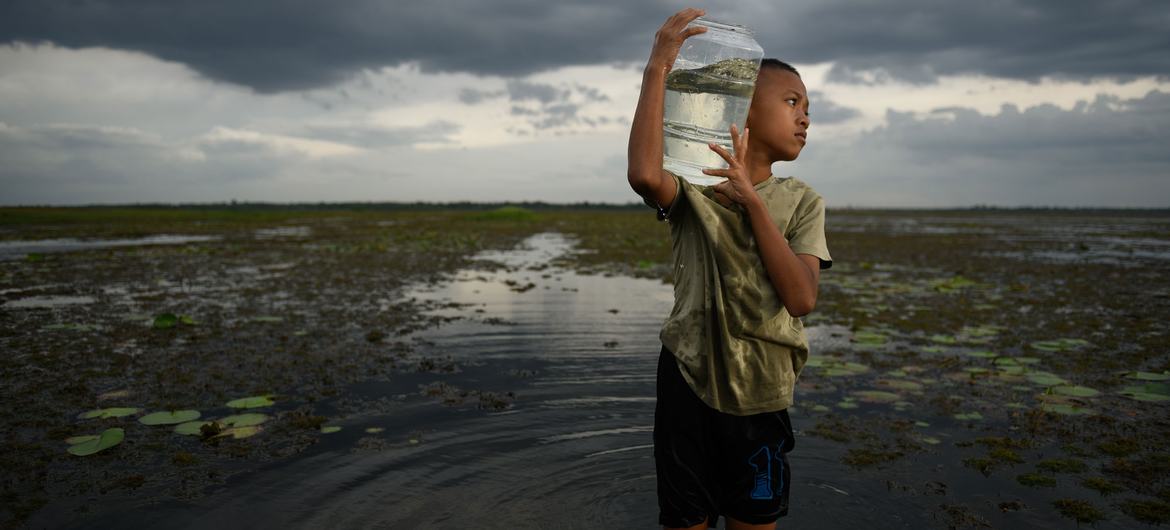 Un niño permanece de pie en un campo inundado, sosteniendo un gran recipiente con agua sobre su hombro.