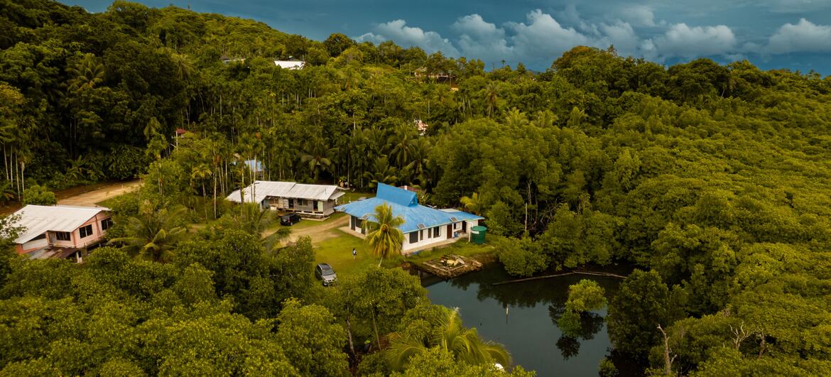 Vista aérea de novos abrigos de desastres em Palau, nestos entre florestas tropicais exuberantes e perto de um corpo de água calma, mostrando a infraestrutura de resiliência climática construída com o apoio da ONU.