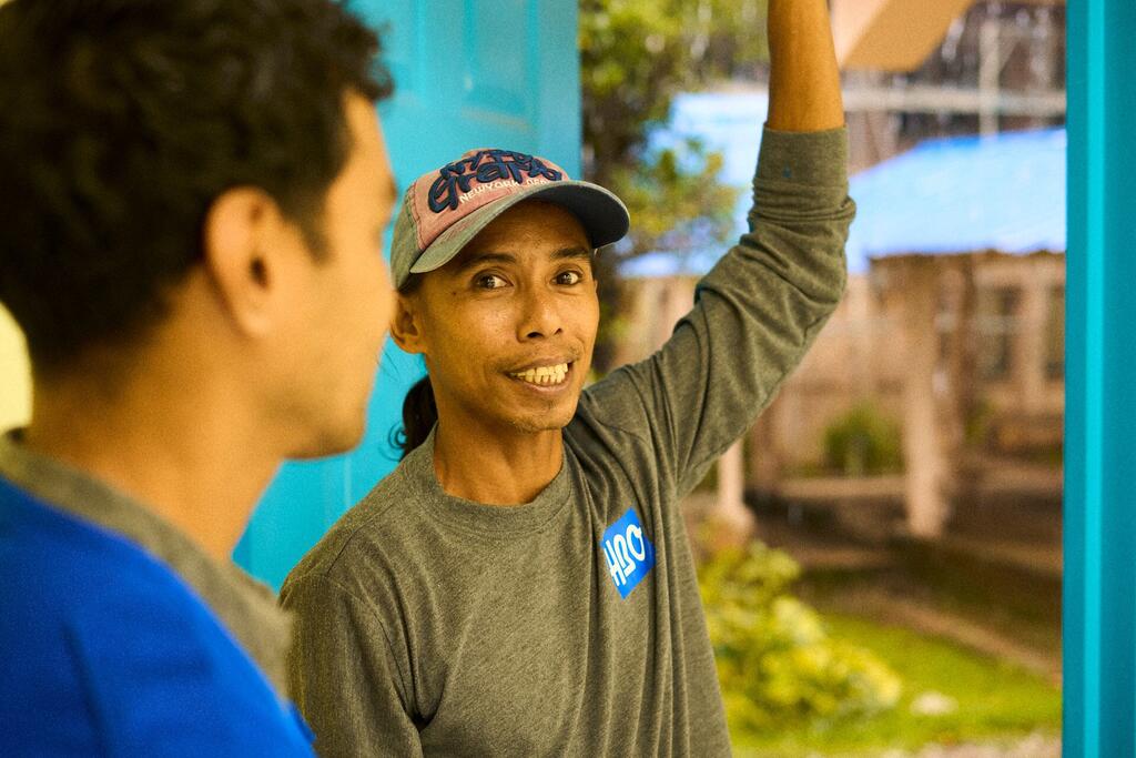 A smiling man in a cap and grey shirt gestures while speaking to another person in Palau, Micronesia, highlighting community engagement on climate change and environmental issues.