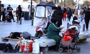 A scene in Beirut, Lebanon showing displaced families living on the streets. In the foreground, a man sleeps on a bench, while a woman in a green tracksuit and hijab sits nearby with a baby in a stroller. Various belongings, including a hookah, are scattered around them. Other people are visible in the background on the city street.