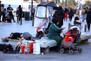Une scène à Beyrouth, au Liban, montrant des familles déplacées vivant dans les rues. En premier plan, un homme dort sur un banc, tandis qu'une femme en combinaison verte et hijab est assise à proximité avec un bébé dans une poussette. Divers biens, y compris un narguilé, sont éparpillés autour d'eux. D'autres personnes sont visibles en arrière-plan dans la rue de la ville.